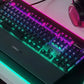 Top-down view of the SteelSeries Apex 5 gaming keyboard with dynamic RGB lighting in purple and green hues, paired with a matching SteelSeries headset on a modern desk setup. Ideal for competitive gaming environments.