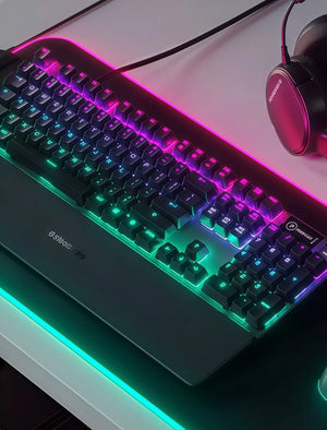 Top-down view of the SteelSeries Apex 5 gaming keyboard with dynamic RGB lighting in purple and green hues, paired with a matching SteelSeries headset on a modern desk setup. Ideal for competitive gaming environments.