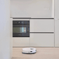A white Ecovacs T30 Pro Omni robot vacuum cleaner operating in a modern kitchen with light-colored hardwood floors. The kitchen features sleek, minimalistic cabinetry and a built-in black and silver Toshiba oven