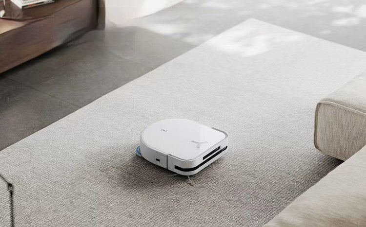 A white Ecovacs Deebot X2 Omni robot vacuum cleaner operating on a light-colored rug in a modern living room. The vacuum is seen cleaning efficiently, with a portion of the sofa and floor visible in the background.