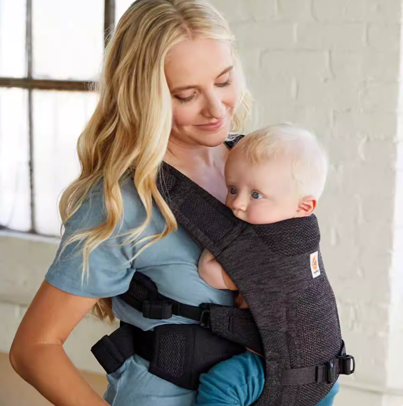 Woman wearing Ergobaby Aerloom baby carrier in dark grey, holding newborn in front-inward position. Lightweight ergonomic baby carrier with breathable mesh fabric, ideal for newborns and infants.