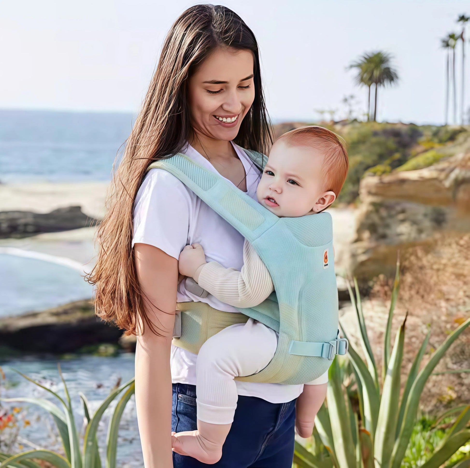Mother using Ergobaby Aerloom baby carrier in light blue, carrying toddler in front-outward position at the beach. Breathable mesh design perfect for summer and outdoor activities.