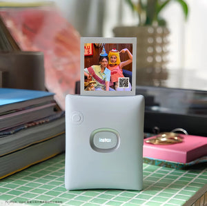 Fujifilm Instax Square Link printer in light blue placed on a tiled desk, printing a fun birthday photo with party hats and QR code. Ideal for home use, gifting, or creative projects.