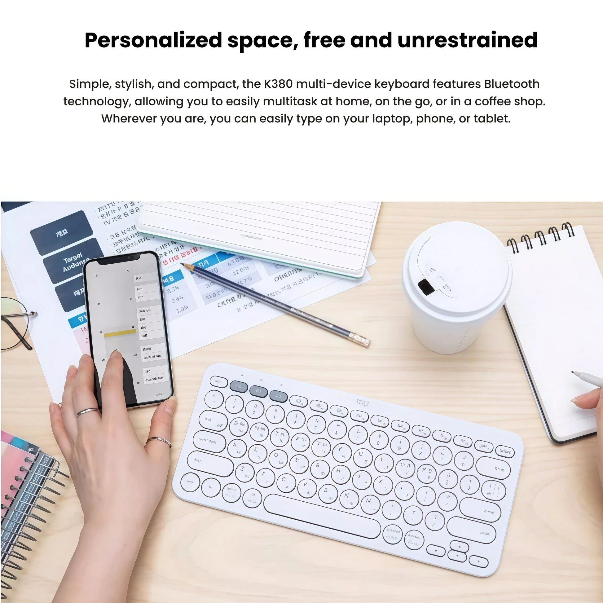 Lifestyle photo of the white Logitech K380 on a wooden desk next to a smartphone, notebook, and coffee cup, illustrating effortless multitasking across laptop, phone, and tablet.