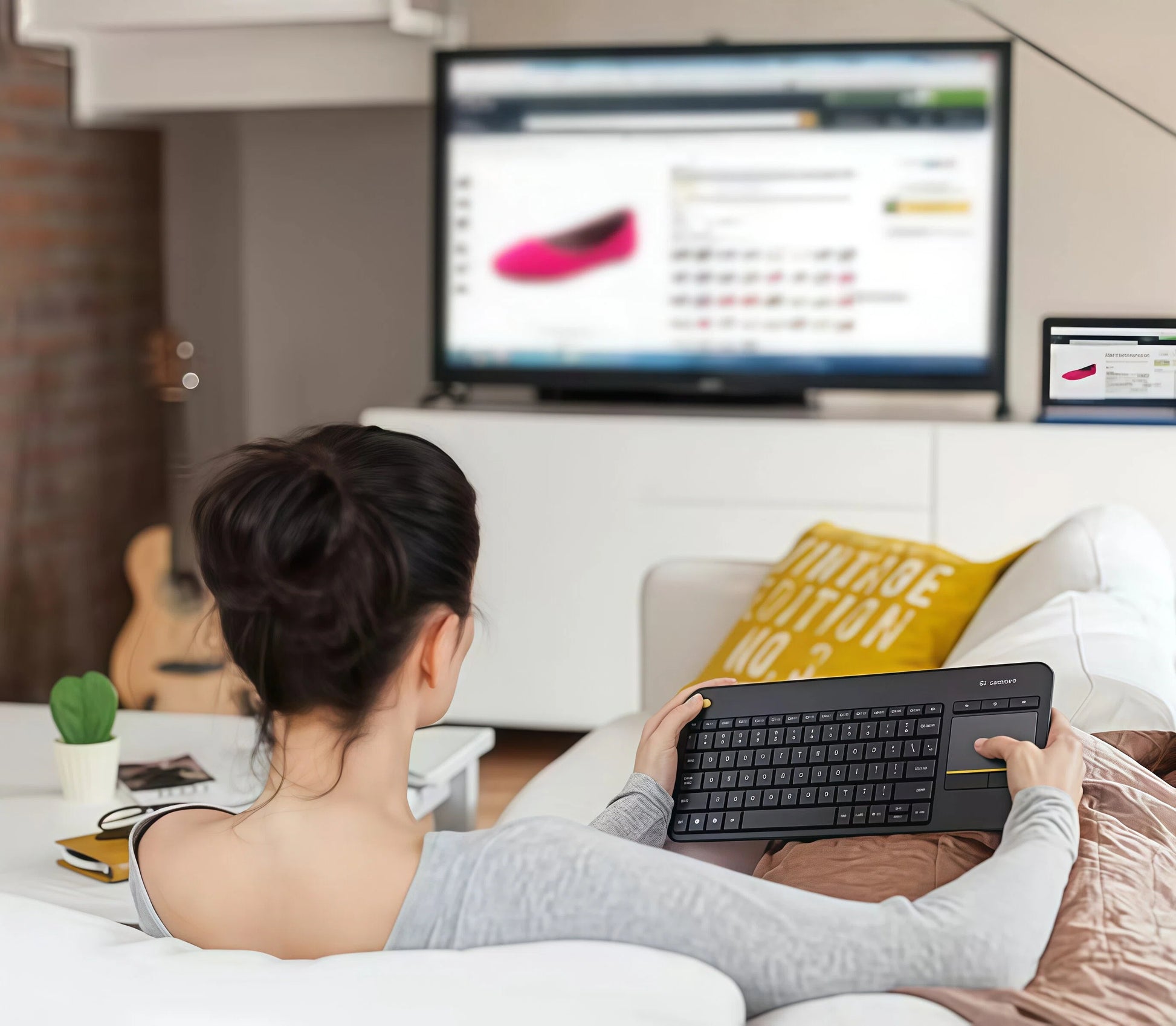 Woman on a sofa using the Logitech K400 Plus to shop online on a big-screen TV, showcasing the keyboard’s convenience for living-room browsing and media control.