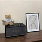 Lifestyle scene with the black Marshall Stanmore III speaker styled on a wooden shelf beside a teddy bear and framed line-art print, blending music and décor.