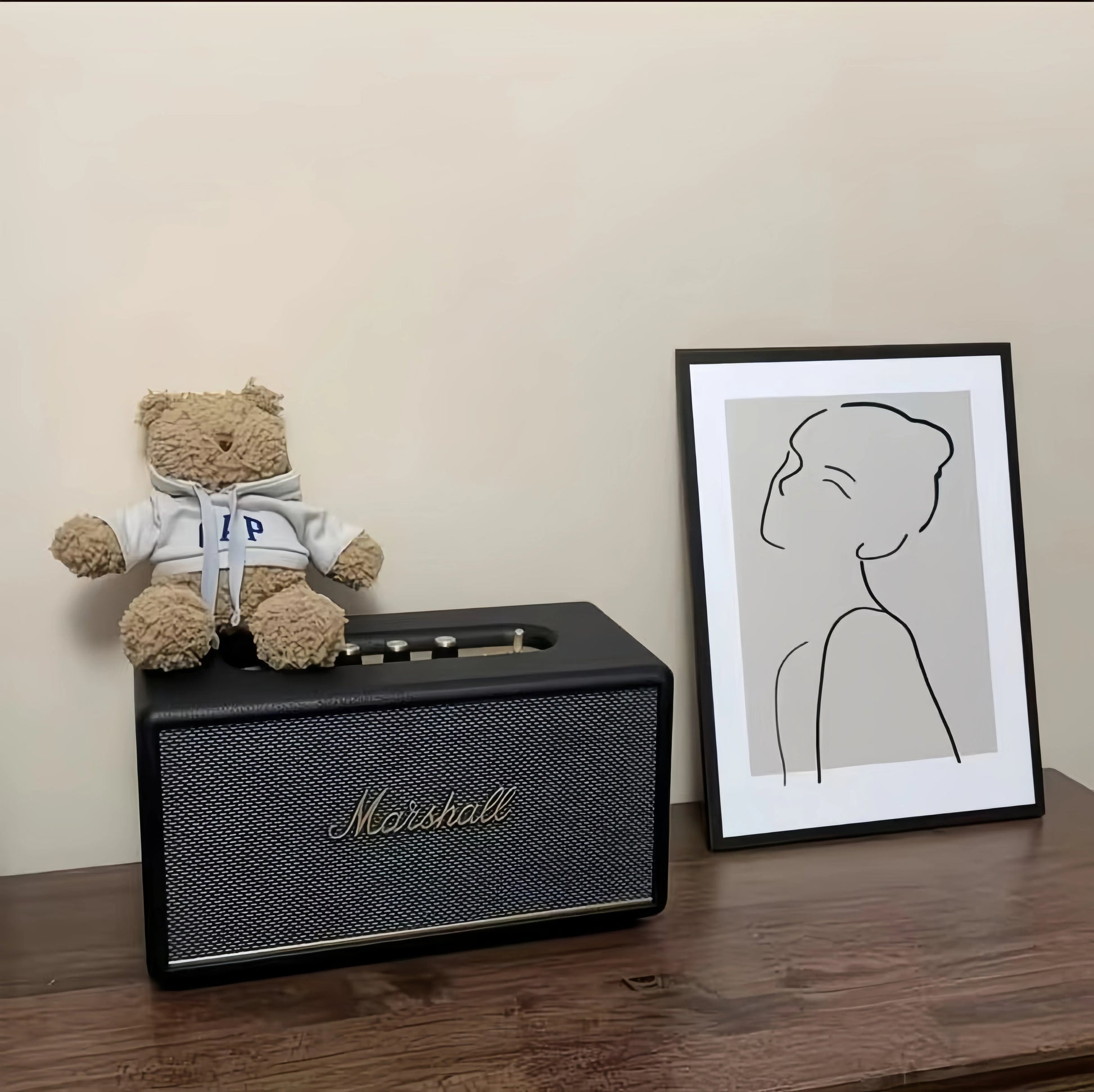 Lifestyle scene with the black Marshall Stanmore III speaker styled on a wooden shelf beside a teddy bear and framed line-art print, blending music and décor.