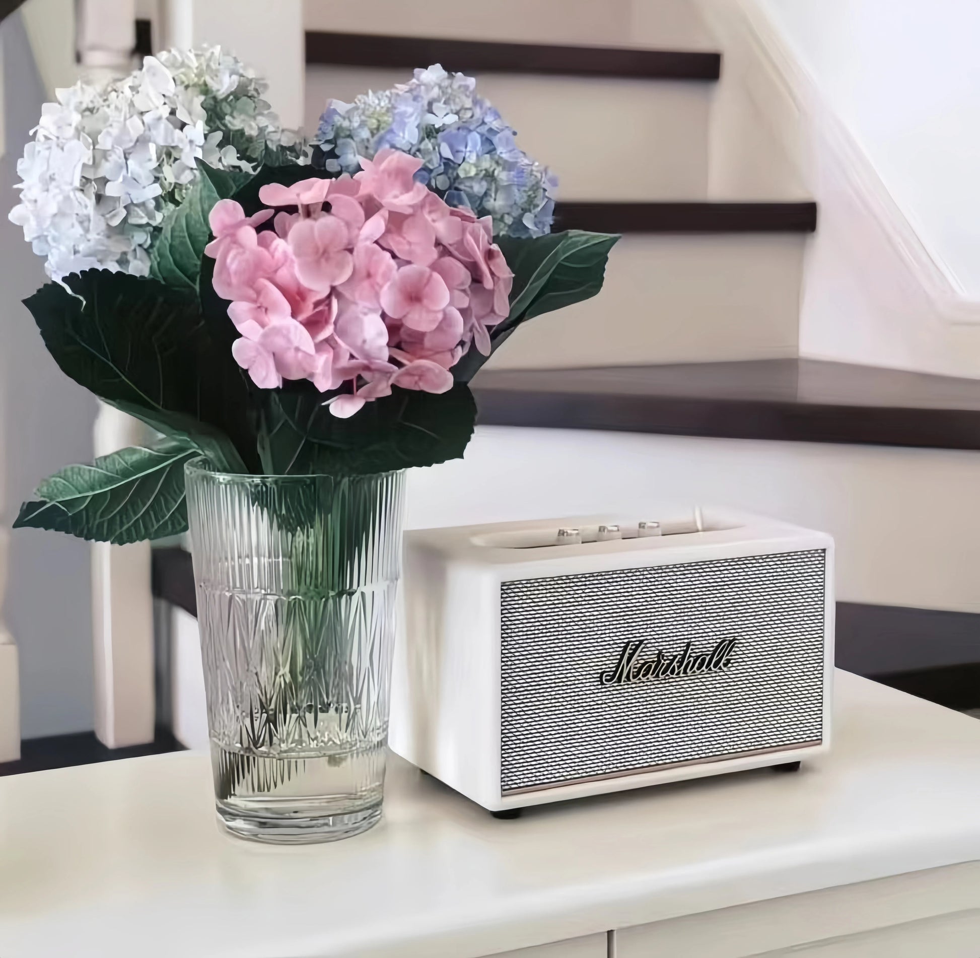  Bright lifestyle shot of the cream Marshall Stanmore III speaker placed next to a vase of pink and blue hydrangeas on a white console table, ideal for home styling inspiration.