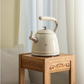 The cream SMEG whistling tea kettle displayed on a light bamboo side table against a bright window, showcasing[1][2] its elegant design as a stylish kitchen decor accessory.