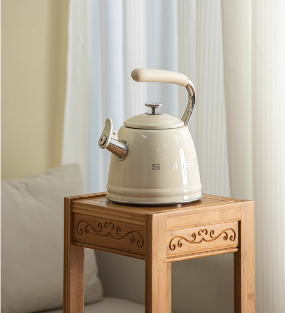 The cream SMEG whistling tea kettle displayed on a light bamboo side table against a bright window, showcasing[1][2] its elegant design as a stylish kitchen decor accessory.