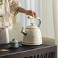 A person holding the handle of a cream SMEG 50's Retro Style Whistling Kettle over a traditional dark wood tea table with a small green teacup, showing its use for daily rituals.