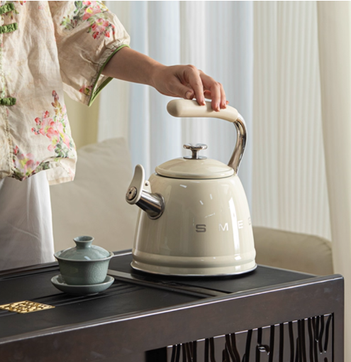 A person holding the handle of a cream SMEG 50's Retro Style Whistling Kettle over a traditional dark wood tea table with a small green teacup, showing its use for daily rituals.