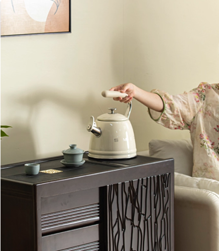 The cream SMEG Retro Stovetop Kettle sitting on a dark wood tea cabinet, ready for pouring, with a person seated on a sofa in the background creating a warm, at-home ambiance.