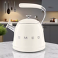 The cream SMEG 2.4 Qt stovetop kettle sitting on a white marble kitchen counter, with a blurred modern kitchen i[2][4][5]n the background, demonstrating its versatile style.