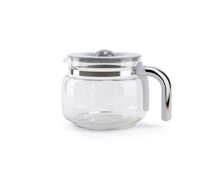 The empty Smeg glass coffee pot sits on a wooden surface surrounded by fresh, whole coffee beans, oranges, and white espresso cups, with steam rising in the background.