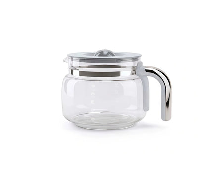 The empty Smeg glass coffee pot sits on a wooden surface surrounded by fresh, whole coffee beans, oranges, and white espresso cups, with steam rising in the background.