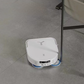 A white Ecovacs Deebot X2 Omni robot vacuum cleaner maneuvering around a metal table leg on a smooth, grey floor. The vacuum is equipped with cleaning brushes and is shown efficiently navigating the space