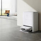A white Ecovacs Deebot X2 Omni robot vacuum cleaner docked in its charging and cleaning station against a wall in a modern home. The station is sleek and minimalistic, with a portion of the living area featuring a chair and large windows visible in the background.