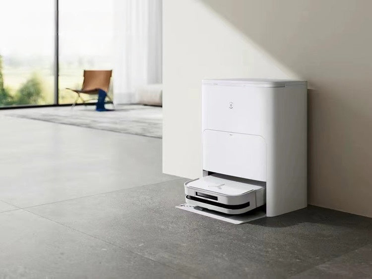 A white Ecovacs Deebot X2 Omni robot vacuum cleaner docked in its charging and cleaning station against a wall in a modern home. The station is sleek and minimalistic, with a portion of the living area featuring a chair and large windows visible in the background.